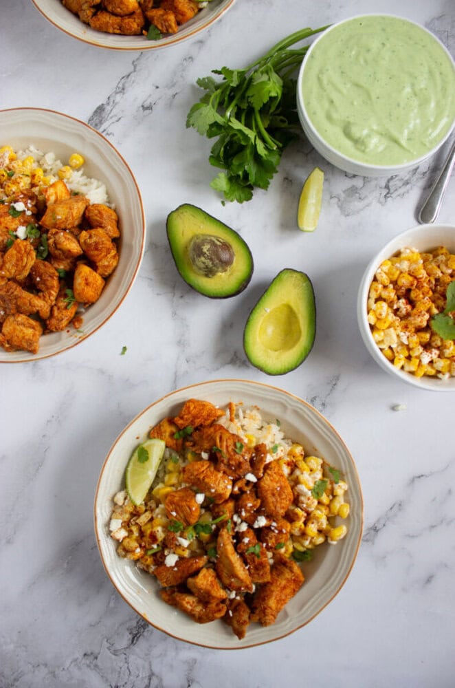 Two bowls of rice topped with seasoned chicken, corn, cheese, and lime. Garnished with cilantro, avocado halves, a bowl of green sauce, and extra corn on a marble surface.