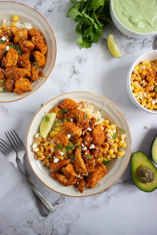 Two bowls of seasoned chicken, corn, and rice are garnished with lime wedges and cheese, surrounded by cilantro, avocado, a bowl of corn, and a bowl of green sauce on a marble surface.