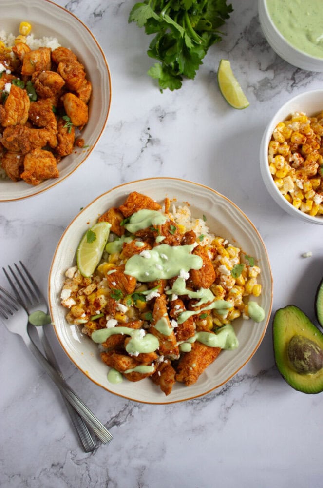 A bowl of rice topped with seasoned chicken, corn, cotija cheese, a lime wedge, and green sauce. Avocado, cilantro, and a bowl of corn are on the side.