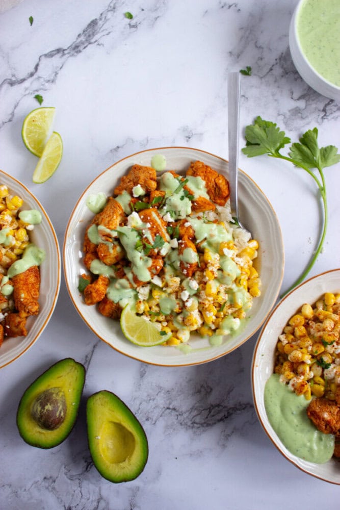 Three bowls of chicken, corn, and sauce are arranged on a marble surface, with lime wedges, fresh cilantro, halved avocado, and a bowl of green dressing nearby.