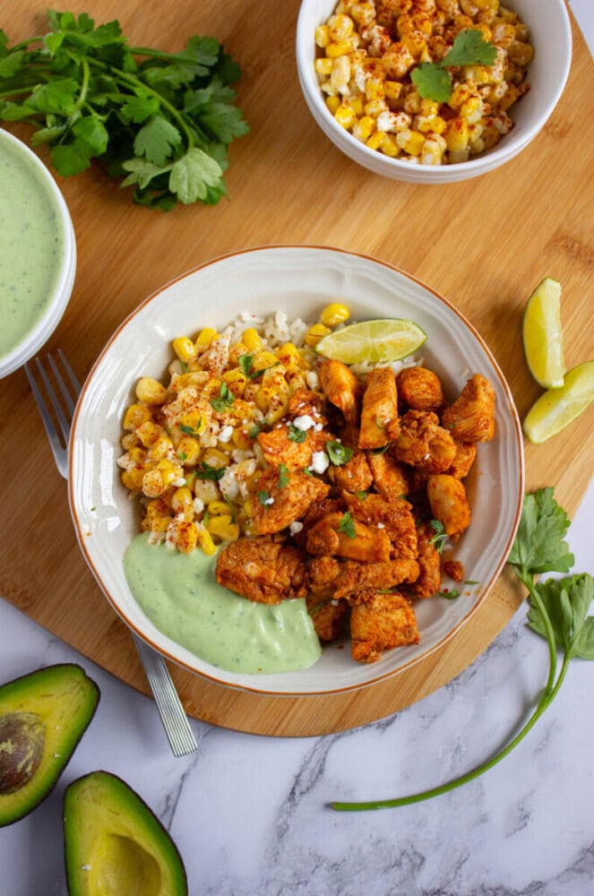 A bowl with seasoned chicken, rice, corn, a wedge of lime, and green sauce, garnished with herbs. A bowl of corn, avocado, lime wedges, and parsley are nearby on a wooden board.