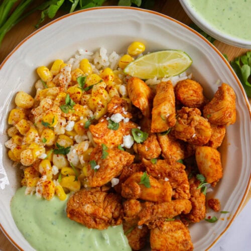A bowl with seasoned chicken pieces, corn kernels, a wedge of lime, and a serving of green sauce, garnished with herbs. Parsley and extra sauce are visible in the background.