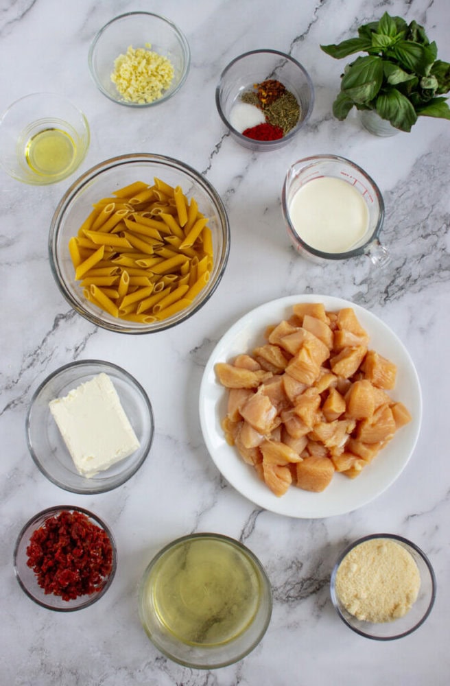 Various ingredients on a marble surface, including uncooked penne pasta, diced raw chicken, cream cheese, sun-dried tomatoes, grated cheese, milk, garlic, oil, seasonings, wine, and fresh basil.