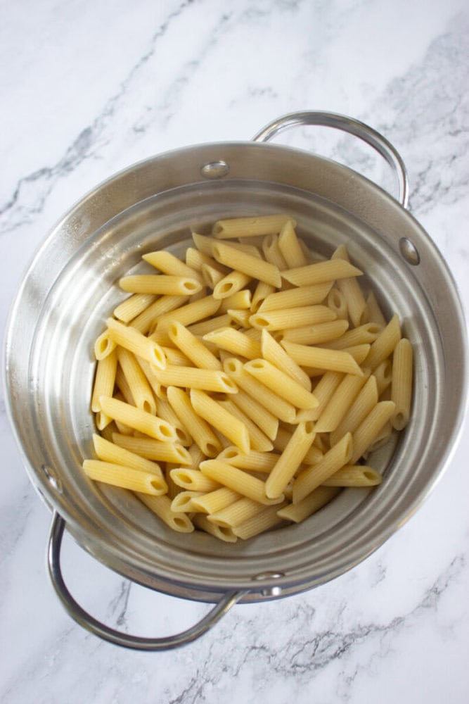 Cooked penne pasta draining in a stainless steel colander over a marble countertop.