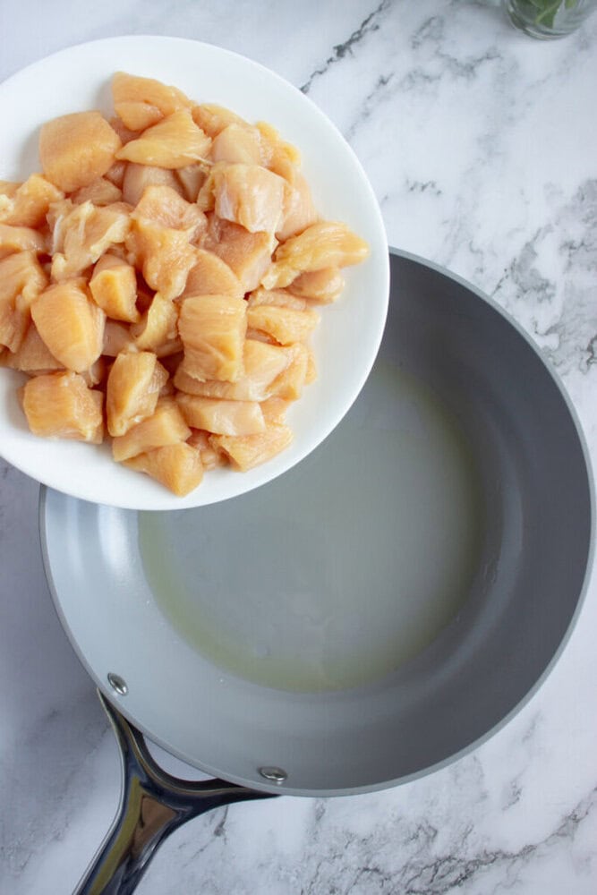 A white plate with raw diced chicken is being poured into a gray frying pan with oil on a marble countertop.