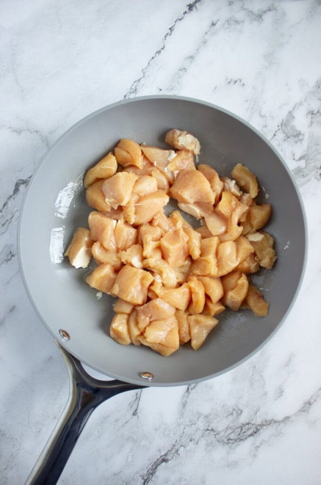 Raw diced chicken pieces in a gray frying pan on a white marble surface.