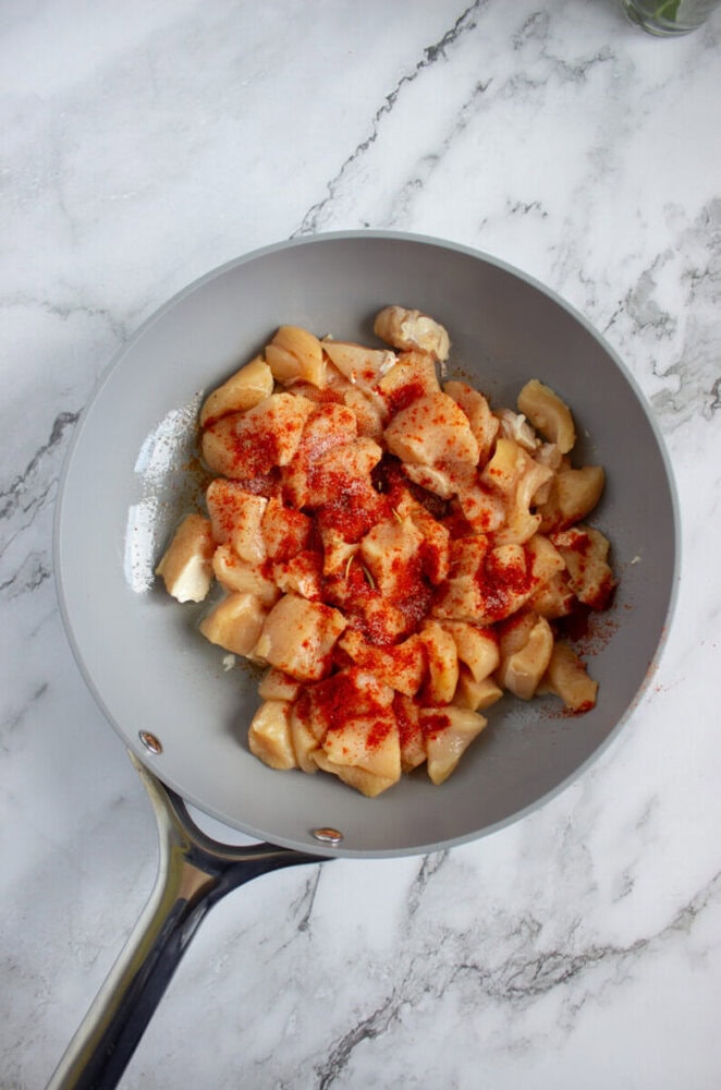 A frying pan with raw, diced chicken pieces sprinkled with red seasoning, placed on a white marble countertop.