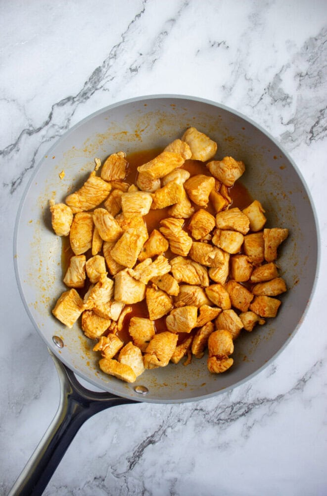 Cooked, seasoned pieces of chicken breast in a frying pan on a marble countertop.
