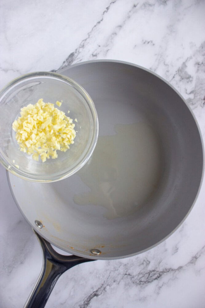 A bowl of minced garlic is held above a pan with oil on a marble countertop.