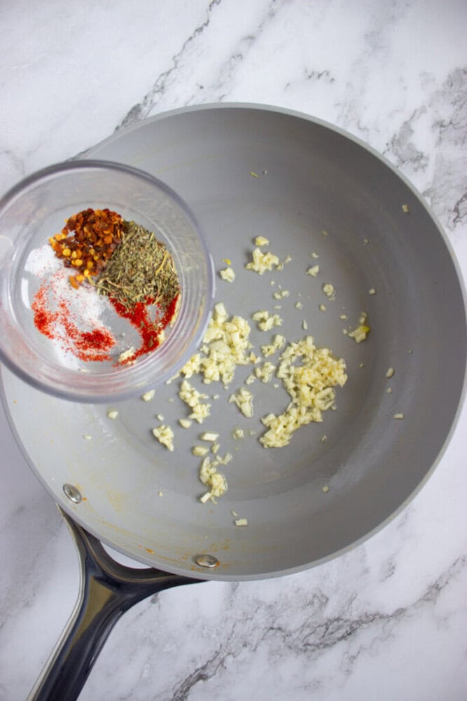 Minced garlic sautéing in a gray skillet, with a small bowl of spices including red pepper flakes, paprika, and dried herbs held above the pan.