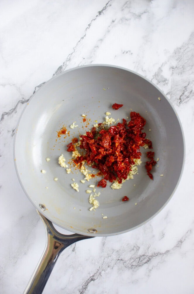 A frying pan contains chopped garlic and sun-dried tomatoes being sautéed on a marble countertop.
