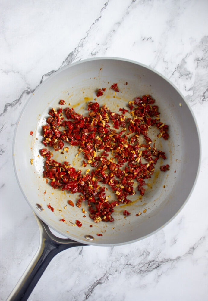 Chopped sun-dried tomatoes and garlic sautéing in a light-colored frying pan on a marble countertop.