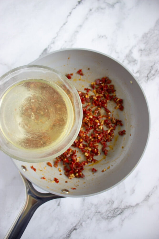 A glass bowl of white wine is being poured into a pan with chopped sun-dried tomatoes on a marble countertop.
