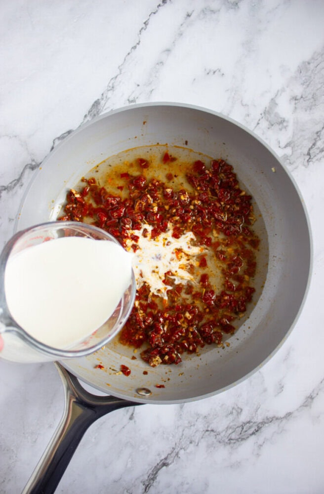A person pours milk from a glass measuring cup into a skillet containing chopped sun-dried tomatoes and oil on a marble countertop.