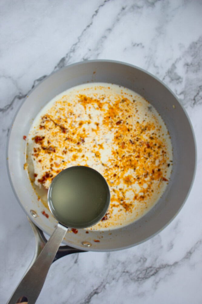 A saucepan with cream and spices on a marble surface, with a metal ladle pouring in a clear liquid, likely lemon juice.