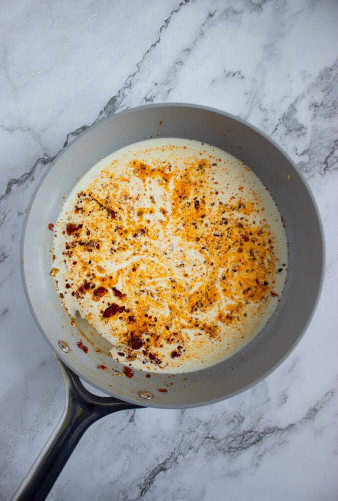 A saucepan with cream, chili flakes, and spices being heated on a marble countertop.