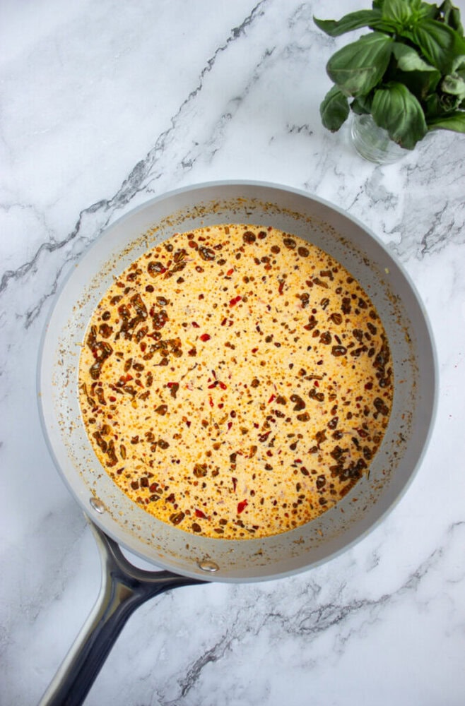 A saucepan containing a creamy, speckled sauce sits on a marble countertop with a bunch of fresh basil nearby.