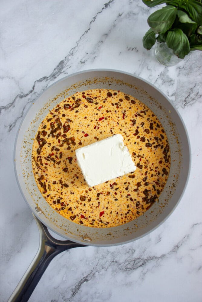 A saucepan with a creamy orange sauce, chili oil, and a block of cream cheese placed in the center, on a marble countertop with basil leaves nearby.