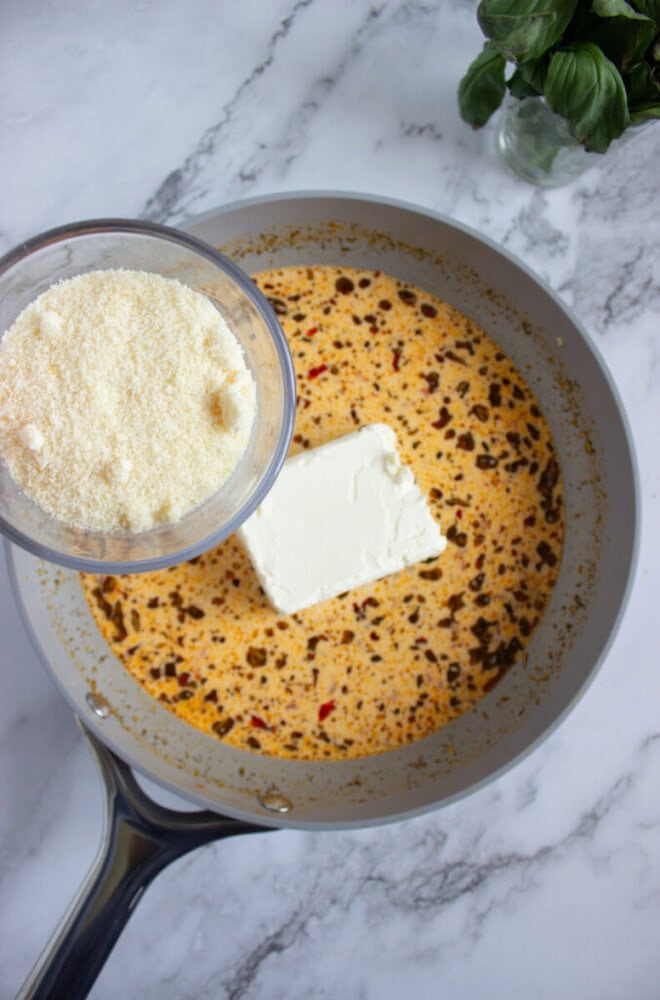 A saucepan with a creamy, spiced sauce and a block of cream cheese; grated parmesan cheese is being added from a measuring cup.
