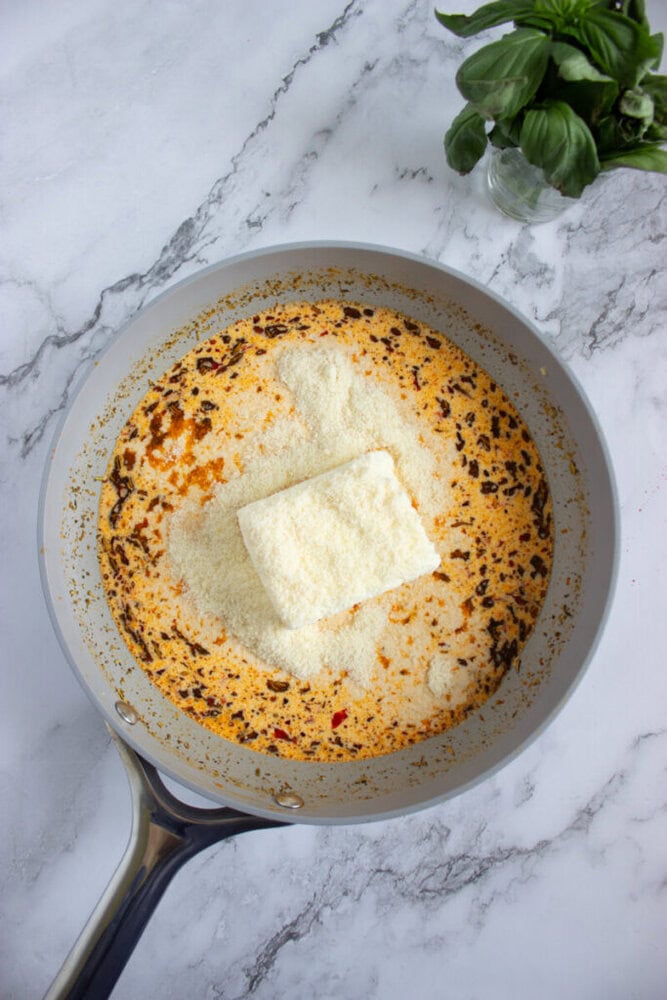 A saucepan with an orange, creamy liquid, a block of cream cheese, and grated cheese on top sits on a marble countertop next to a small bunch of fresh basil.