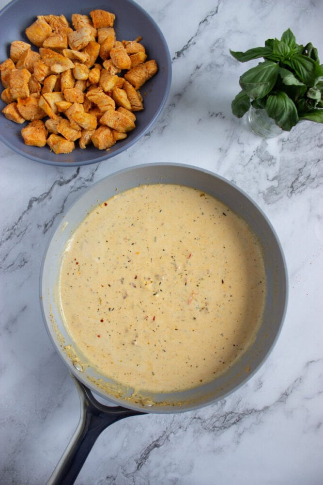 A frying pan with creamy sauce sits on a marble surface next to a bowl of cooked, seasoned chicken pieces and a small vase of fresh basil.