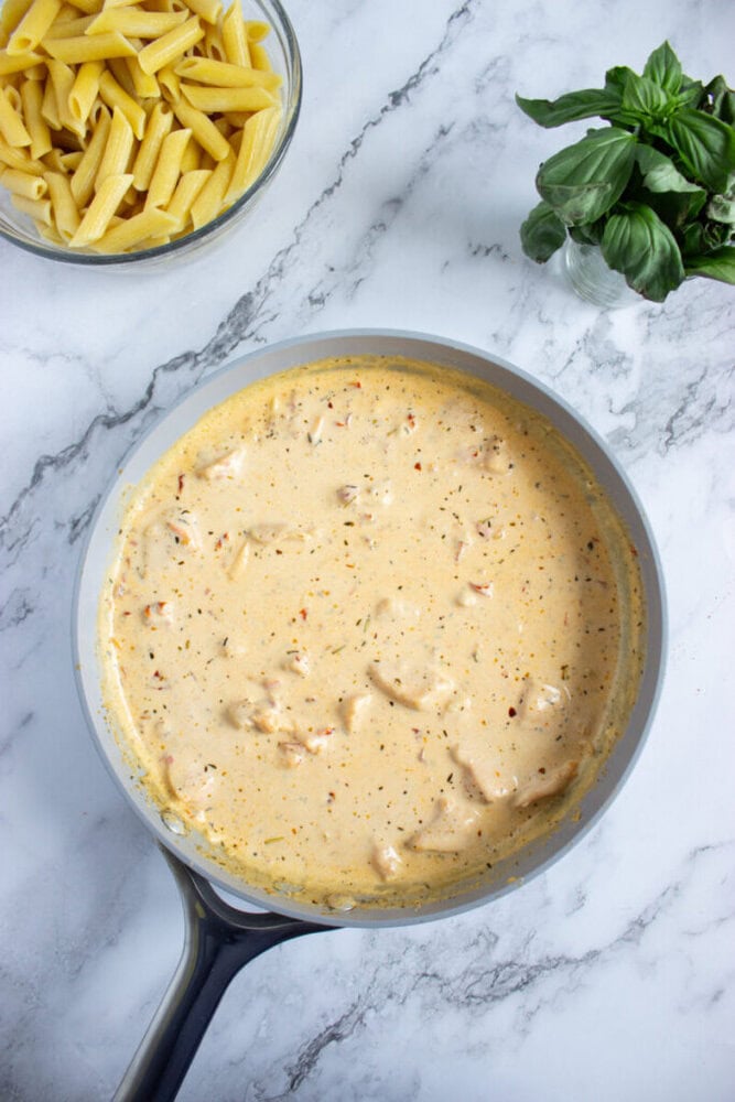 A pan of creamy sauce with chicken pieces sits on a marble counter next to a bowl of uncooked penne pasta and a bunch of fresh basil.