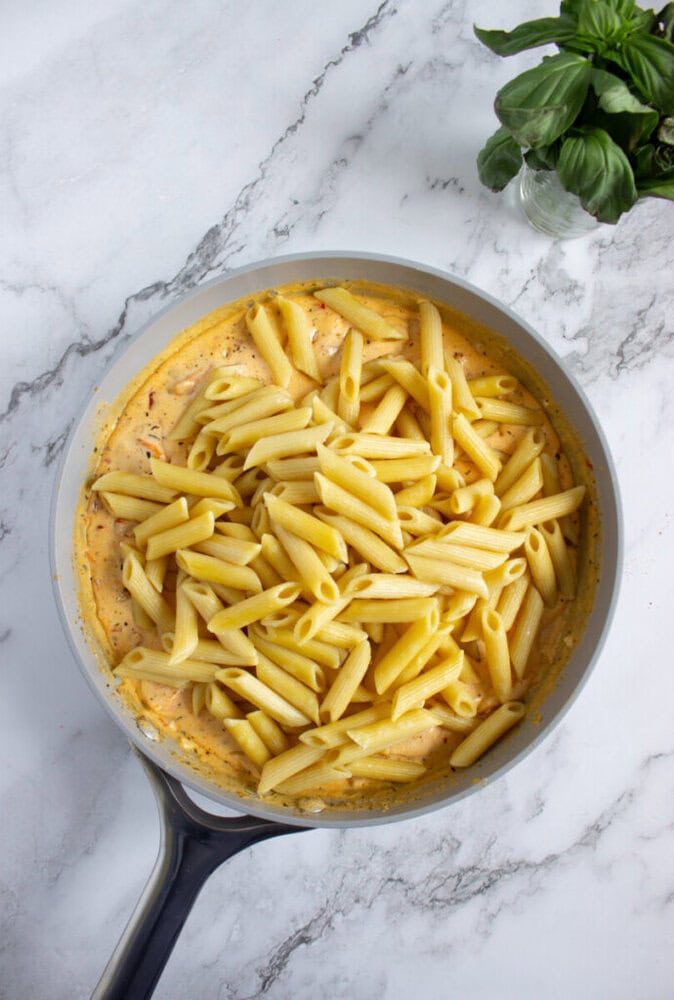 Cooked penne pasta sits on top of a creamy orange sauce in a frying pan, placed on a marble countertop with a small basil plant nearby.