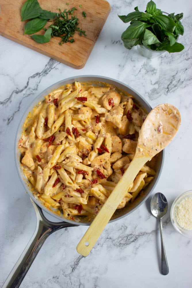 A skillet of creamy penne pasta with chicken and sun-dried tomatoes sits on a marble counter, surrounded by fresh basil, chopped herbs, parmesan, and utensils.