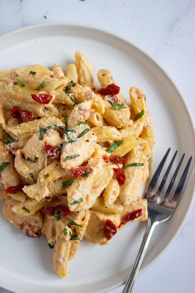 A plate of creamy penne pasta with pieces of chicken, sun-dried tomatoes, parmesan, and chopped herbs. A fork rests on the side of the plate.
