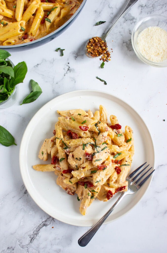 A plate of creamy penne pasta with chicken, sun-dried tomatoes, and fresh basil, served with a fork; grated cheese and red pepper flakes are on the side.