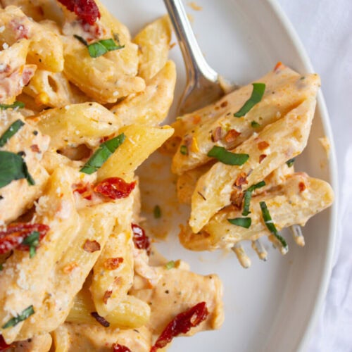 A plate of creamy penne pasta with sun-dried tomatoes and fresh herbs, with a fork holding a bite-sized portion.