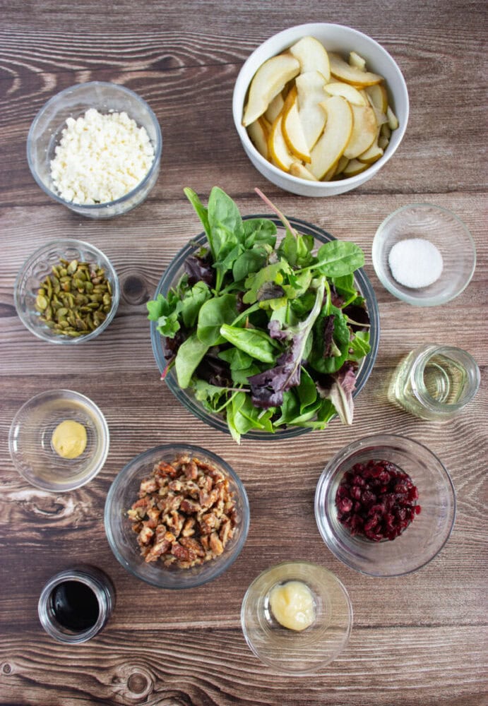 Overhead view of salad ingredients in bowls, including mixed greens, sliced pears, crumbled cheese, nuts, seeds, dried cranberries, and various dressings, arranged on a wooden surface.