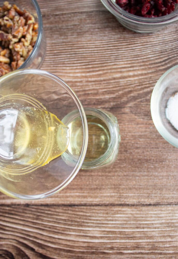 Overhead view of honey being poured into a small glass jar, with bowls of walnuts, dried cranberries, and salt on a wooden surface.