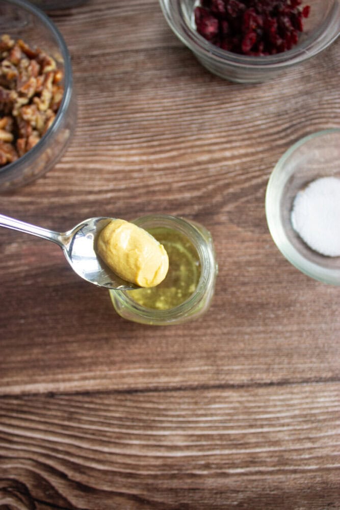 A spoon with a dollop of mustard is held above a small jar of oil, with bowls of nuts, dried cranberries, and salt nearby on a wooden surface.