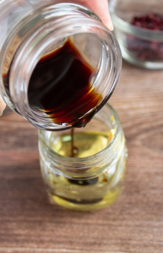 A hand pours dark soy sauce from a glass jar into another jar containing olive oil, on a wooden surface.