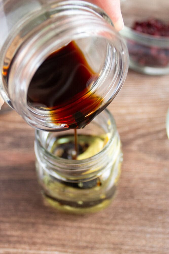 A jar of dark liquid, likely soy sauce, being poured into a small glass jar containing other ingredients on a wooden surface.