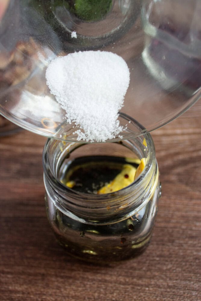 A bowl of salt is being poured into a glass jar containing oil and spices on a wooden surface.