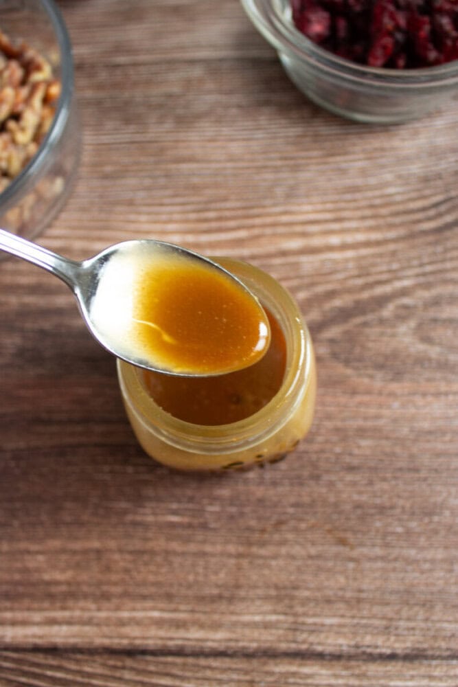 A spoon holds brown liquid above a small glass jar, with bowls of nuts and dried cranberries visible in the background on a wooden surface.