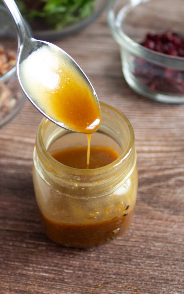 A spoon drizzles brown vinaigrette dressing into a small glass jar, with bowls of salad ingredients in the background on a wooden surface.