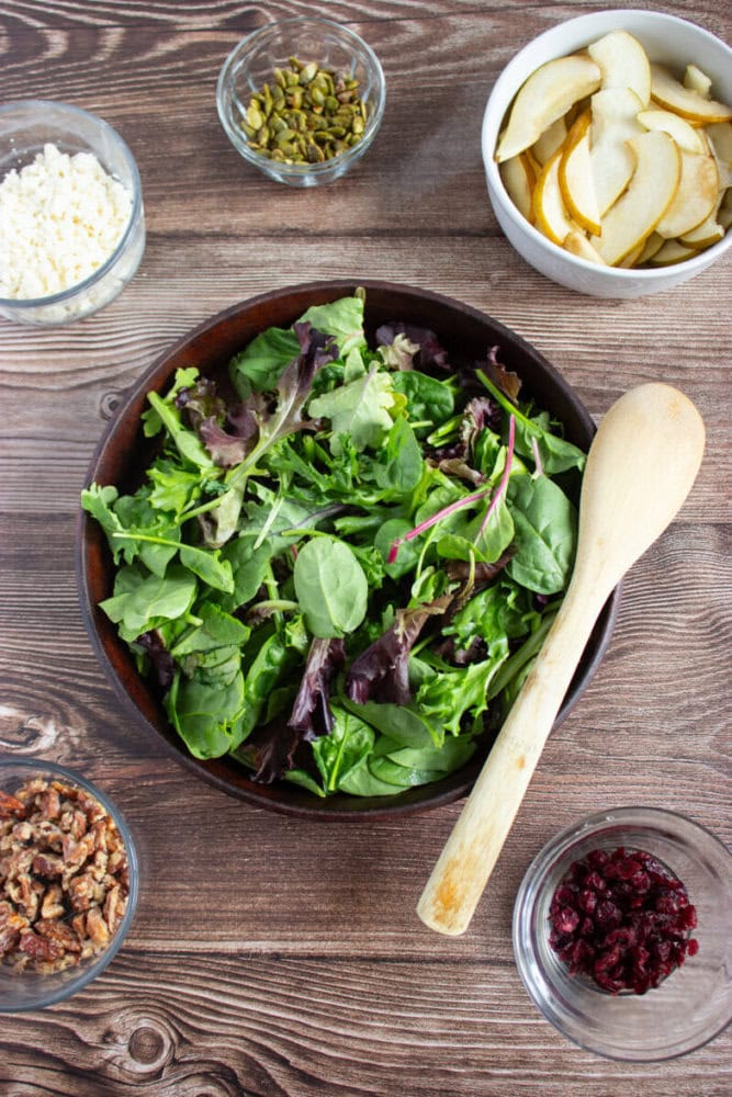 A bowl of mixed salad greens with a wooden spoon, surrounded by small bowls of sliced pears, feta cheese, pistachios, pecans, and dried cranberries on a wooden surface.