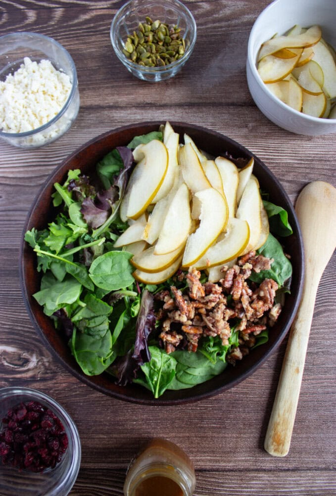 A bowl of mixed greens topped with sliced pears and candied pecans, surrounded by bowls of feta cheese, pumpkin seeds, dried cranberries, and dressing on a wooden surface.