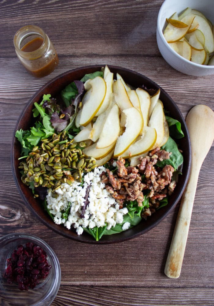 A salad with mixed greens, sliced pears, crumbled cheese, candied nuts, and seeds in a bowl. Surrounding items include dressing, dried cranberries, a bowl of pear slices, and a wooden spoon.