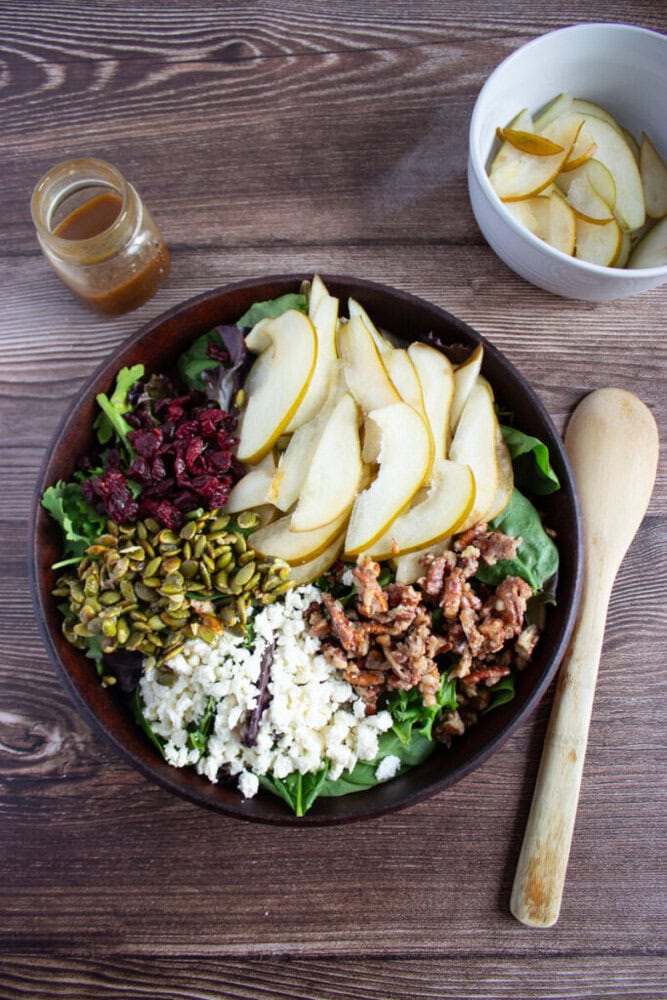 A salad with sliced pears, feta cheese, candied walnuts, dried cranberries, and pumpkin seeds on greens, with a small jar of dressing and a bowl of sliced pears nearby.
