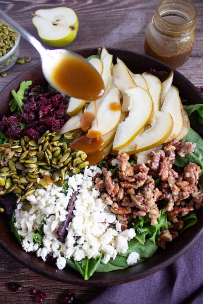 A bowl of salad with sliced pears, crumbled cheese, candied walnuts, dried cranberries, and pumpkin seeds, with dressing being poured from a spoon.