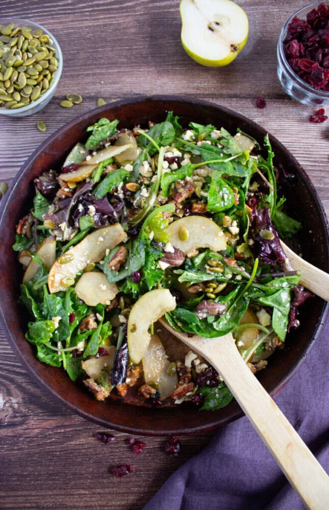 A bowl of mixed green salad with sliced pears, pecans, pumpkin seeds, dried cranberries, and feta cheese, served with wooden salad servers on a wooden table.