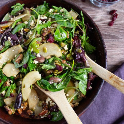 A bowl of mixed green salad with sliced pears, dried cranberries, nuts, seeds, and feta cheese, with wooden salad servers beside a glass of dried cranberries and half a pear.