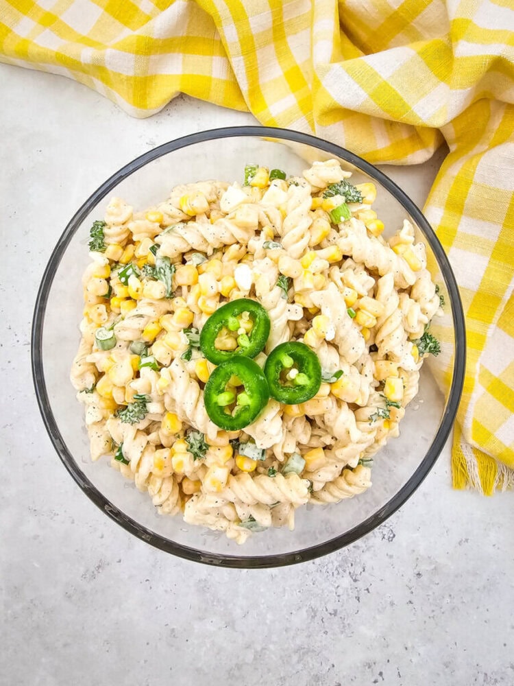 A bowl of creamy pasta salad with corn, broccoli, and sliced jalapeños on top, placed on a light gray surface near a yellow checkered cloth.