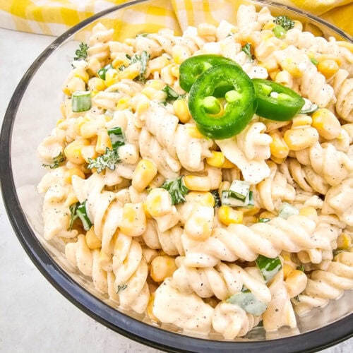 A glass bowl filled with creamy pasta salad, featuring spiral noodles, corn, green onions, and garnished with sliced jalapeño. A yellow checkered cloth in the background.