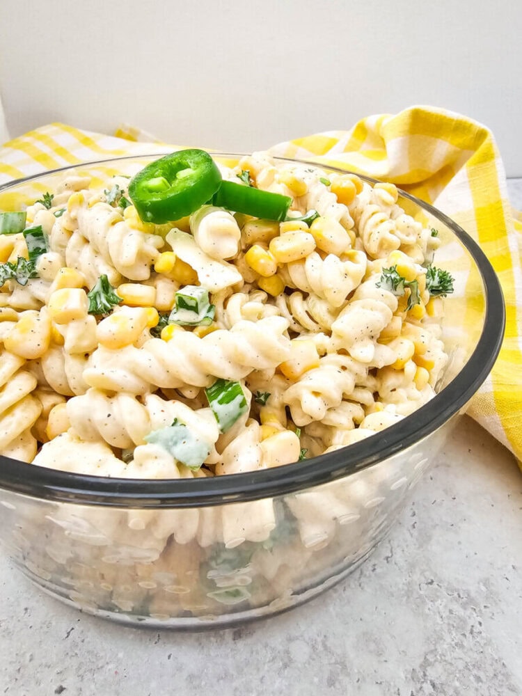A glass bowl filled with creamy pasta salad, featuring fusilli, corn, chopped green peppers, and herbs, placed on a gray surface with a yellow-checked cloth in the background.