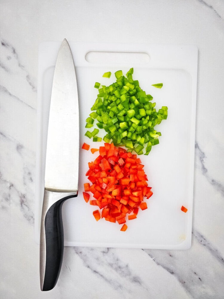 Chopped green and red bell peppers on a white cutting board with a large kitchen knife to the left.